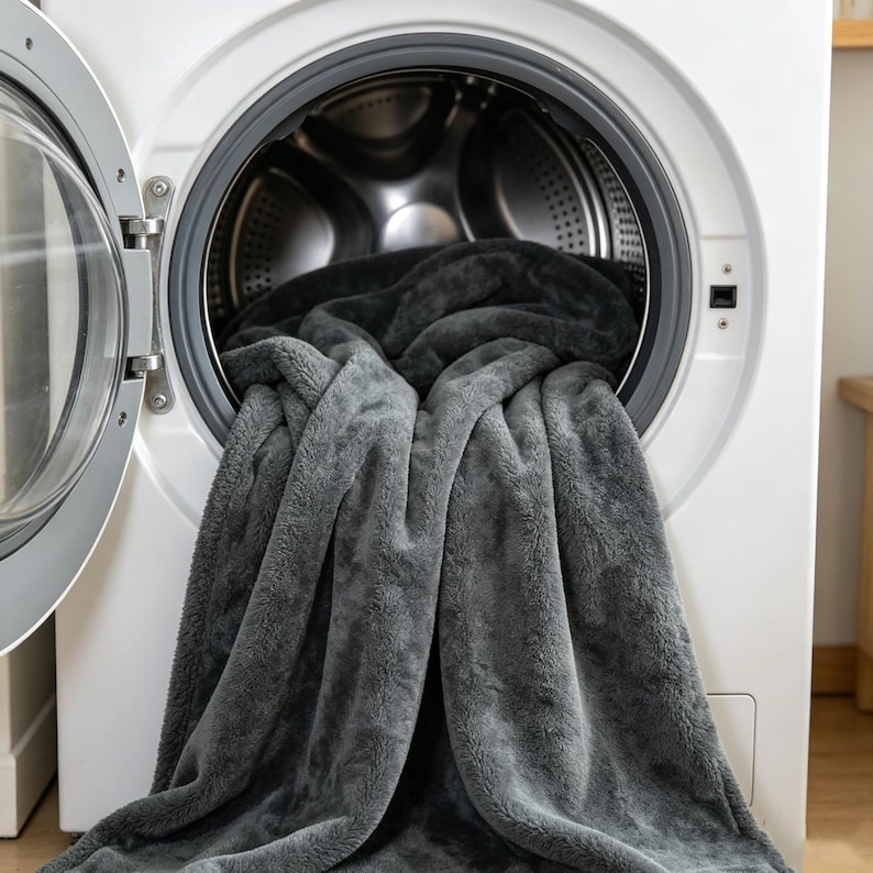 May include: A gray plush blanket partially inside a white washing machine. The blanket is a soft, textured material, with a darker gray color inside the machine. The washing machine door is open, revealing the interior.