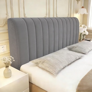 May include: A tall, rectangular headboard upholstered in smooth, medium-gray fabric featuring vertical channel tufting. The headboard stands behind a bed dressed in white linens with two rectangular pillows featuring a fine, light-gray striped pattern.