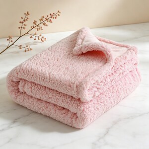 May include: A folded, light pink sherpa blanket. The blanket is soft and fluffy, with a textured surface. The edges are neatly folded, and the blanket appears to be made of a plush material. The blanket is folded in thirds, ready for use.