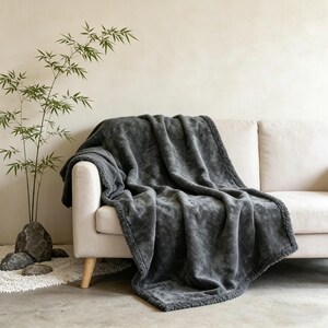 May include: A dark gray plush throw blanket draped over a cream-colored sofa. The blanket has a soft, textured appearance and a contrasting border. A bamboo plant and decorative stones are in the background.