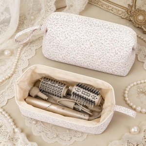 Può includere: Una trousse rettangolare in tessuto chiaro con un piccolo motivo floreale rosa e verde. La borsa è dotata di chiusura a zip e manico laterale. All'interno è visibile un dispositivo per lo styling dei capelli beige con vari accessori.