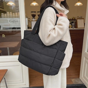 May include: A black quilted tote bag with a rectangular shape, horizontal stitching, and two shoulder straps. The bag features a smooth, padded fabric construction with side pockets and a small rectangular brand tag on the lower right side.