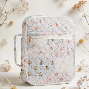 May include: A rectangular quilted carrying case featuring a patchwork design of floral patterns in shades of light blue, pink, and white. The case includes a side handle, a gold-toned zip closure, and a front zipped pocket with a gold pull.