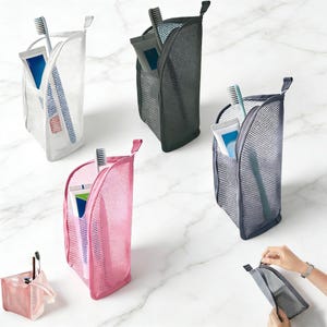 May include: A collection of upright mesh toiletry bags in white, black, pink, and blue, each containing a toothbrush and a tube of toothpaste. The bags feature a zipped opening and a small loop at the top for hanging, designed for travel storage.
