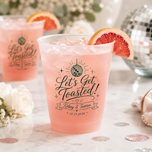 Può includere: Un bicchiere di plastica trasparente riempito con una bevanda rosa, ghiaccio e una fetta di pompelmo. Il bicchiere presenta una scritta verde scuro "Let's Get Toasted!", "Kelsey & Taverin" e la data "12.19.2026" con un grafico a palla da discoteca.