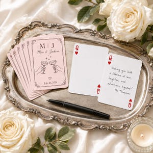 May include: A stack of light pink playing cards featuring a line art illustration of two hands toasting with champagne glasses, the initials M | J, the names Mia & Jackson, and the date July 26, 2026, alongside a black pen and two white cards.