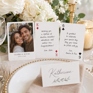 May include: Three white rectangular cards designed like playing cards. One features a photo of a couple with the text CLAIRE & LUCAS JULY 19, 2025. The others contain printed messages and signatures. A white place card with the name Katherine is in front.