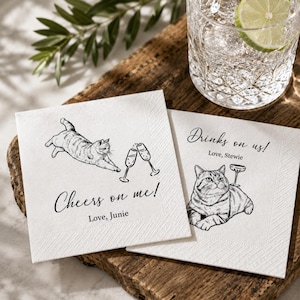 May include: Two white square paper napkins featuring black ink illustrations of cats and champagne glasses. One napkin displays a cat stretching with the text Cheers on me! Love, Junie. The other shows a cat lying down with the text Drinks on us! Love, Stewie.