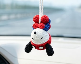 Op de afbeelding: Een gehaakte hangende decoratie aan een wit koord, met een figuur met een witte kop, zwarte oren en een rood en blauw lichaam. Het figuurtje hangt ondersteboven en heeft een ronde vorm met een textuur van rood en blauw garen.
