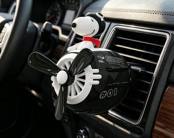 Op de afbeelding: Een zwart-witte auto-luchtverfrisser in de vorm van een vintage vliegtuig met een Snoopy-figuur met een pilotenmuts en een rode sjaal. De propeller is zwart-wit en op de zijkant van het vliegtuig staat de tekst Snoopy en het nummer 01.