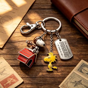 May include: A metal keychain featuring a Snoopy figure in an aviator cap sitting on a red doghouse, a yellow Woodstock charm, and a silver rectangular tag engraved with 75TH ANNIVERSARY PEANUTS - 75 YEARS OF JOY - attached to a split ring.