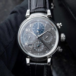 Peut inclure: Une montre de luxe avec un cadran gris, des aiguilles argentées et un bracelet en cuir noir. La montre comporte plusieurs sous-cadrans, dont un indicateur de phase de lune, et la marque "IWC" est visible. Un accessoire sophistiqué.