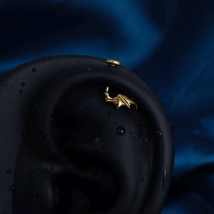 May include: A gold-toned ear cartilage stud featuring a small, detailed bat wing design. The jewelry is secured in a black ear-shaped display model, with a flat circular backing visible at the top of the ear cartilage.