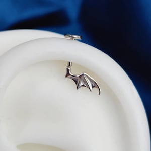 May include: A silver-toned metal body jewelry piece featuring a small, detailed bat wing design. The jewelry has a flat circular backing and a straight post, positioned securely against a white, curved, ear-shaped display form.