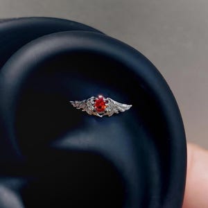 May include: A silver-toned body jewelry piece featuring a central oval-shaped red gemstone flanked by detailed metallic wings. The ornament is designed with a prong setting for the stone and intricate feather textures on the wing-shaped metal accents.