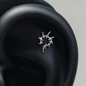 Puede incluir: Una pieza de joyería para piercing de oreja en tono plateado con un diseño circular de púas y protuberancias afiladas en forma de espinas que se extienden hacia afuera. El metal tiene un acabado pulido y reflectante en forma de espiral abierta.
