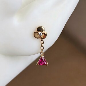 May include: A gold stud earring featuring a three-petaled flower design with a small clear center stone. A delicate gold chain hangs from the flower, connecting to a triangular, faceted, deep pink gemstone pendant at the bottom of the earring.