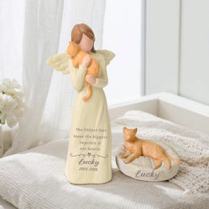 May include: A cream-colored angel figurine holding an orange cat, with the text "The littlest feet leave the biggest imprints in our hearts," "Lucky," and "2005-2026." Beside it is a separate orange cat figurine on a base labeled "Lucky."