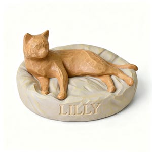 May include: A tan cat figurine resting on a circular, textured off-white base. The cat is lying down with its head turned to the left. The name LILLY is carved in capital letters into the front of the base, which has a carved, uneven surface.
