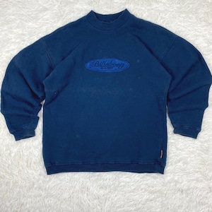 Può includere: Una felpa a maniche lunghe blu scuro con collo alto e polsini a coste. Una toppa ovale è centrata sul petto con la parola Billabong ricamata in filo blu. Una piccola etichetta rettangolare del marchio è visibile sul lato inferiore.