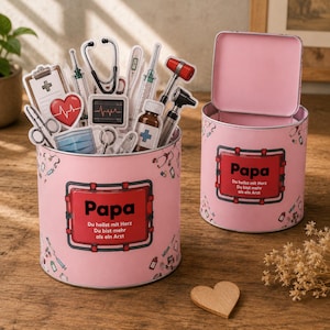 Puede incluir: Dos recipientes de metal rosa con pegatinas de temática médica en el interior. Los recipientes tienen un rectángulo rojo con la palabra "Papa" y texto en alemán. La tapa de un recipiente está abierta. Un corazón de madera está en la superficie.