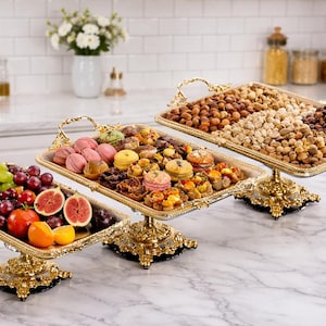 May include: Three gold-coloured serving platters, each laden with food. One holds fresh fruit, including grapes and figs. Another contains pastries, and the third is filled with nuts and dried fruit. The trays are supported by ornate, decorative stands.