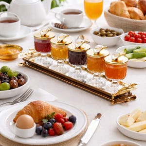 May include: A breakfast table setting with various foods and drinks. Includes a plate with a croissant, berries, and an egg. There are small jars of preserves, tea, juice, bread, olives, and cheese. The table is set with white dishes and a white tablecloth.