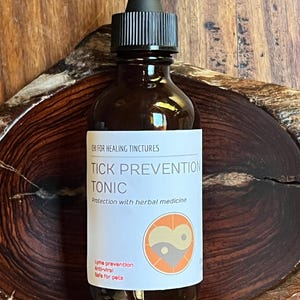 Puede incluir: Una botella de vidrio marrón con un cuentagotas negro y una etiqueta blanca. La etiqueta dice "TICK PREVENTION TONIC" e incluye el texto "Protección con medicina herbal". La botella es para la prevención de Lyme y es segura para mascotas.
