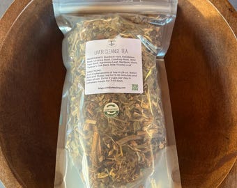 May include: A clear plastic bag filled with dried herbs, labeled "LIVER CLEANSE TEA," sits in a wooden bowl. The label lists ingredients like burdock root and dandelion root. The bag is sealed and includes directions for use.