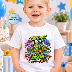 May include: A white t-shirt featuring a graphic of four Teenage Mutant Ninja Turtles with the text "BIRTHDAY BOY!", "JASON", "DUDE! IT'S MY 5TH.", and action words "SMASH!", "BAM!", "SLASH!", and "ATTACK!" surrounded by colourful paint splatters.