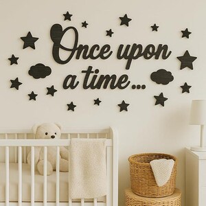 May include: Black wooden wall decor with the phrase "Once upon a time..." and star and cloud shapes. The decor is above a white crib with a teddy bear and a woven basket.