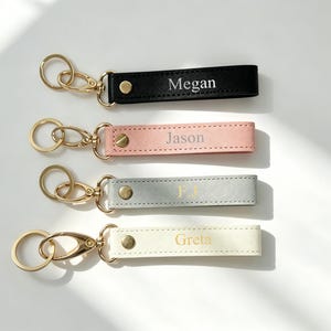 May include: Four personalised keychains in black, pink, grey, and cream. Each keychain has a gold-coloured clasp and a strap with a name printed in gold. The names are Megan, Jason, P.J., and Greta.