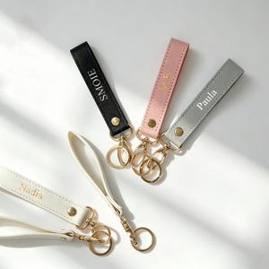 Puede incluir: Cuatro llaveros de cuero en negro, rosa, gris y crema. Cada llavero tiene un cierre dorado y una correa con un nombre impreso: "Smoie", "Nadia" y "Paula". Los llaveros están sobre una superficie blanca.