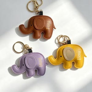 Könnte beinhalten: Drei Leder-Elefanten-Schlüsselanhänger in Braun, Lila und Gelb. Jeder Schlüsselanhänger hat einen goldfarbenen Ring und einen Namen auf der Seite. Der braune Elefant hat den Namen "Fiona", der lila "Hazel" und der gelbe "Elena".