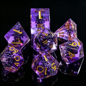 Peut inclure: Un ensemble de sept dés polyédriques violets translucides avec des chiffres dorés. Chaque dé contient une inclusion sombre, texturée et d'aspect organique suspendue dans la résine. L'ensemble comprend diverses formes pour les jeux.
