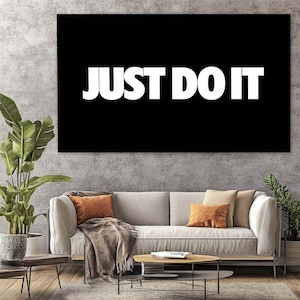 Peut inclure: Une grande toile noire avec le texte blanc "JUST DO IT". L'œuvre est accrochée au-dessus d'un canapé avec des coussins orange et beiges. Une table basse en bois et des plantes en pot complètent la décoration de la pièce.