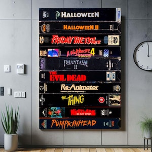 Puede incluir: Una obra de arte montada en la pared con una colección de cintas VHS vintage. Los títulos incluyen "Halloween", "Viernes 13 Parte 2" y "The Evil Dead". Las cintas están apiladas verticalmente, creando un tema de película de terror retro.