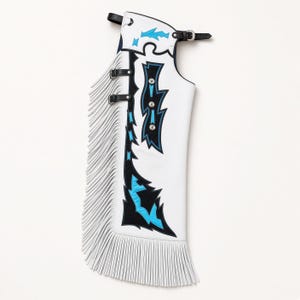 May include: White leather chaps featuring long white fringe along the side. The design includes black and bright blue jagged geometric overlays, silver-toned metal studs, and black adjustable buckle straps at the waist and thigh area.