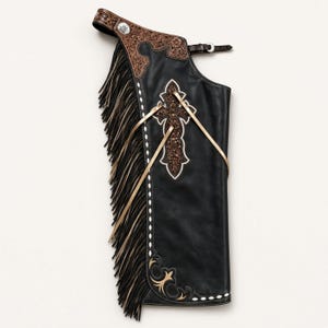 May include: A black leather chap featuring a brown floral-tooled leather yoke, long black and tan fringe, and a central cross applique with intricate tooling. The piece includes a buckle strap at the top and white decorative stitching along the edge.