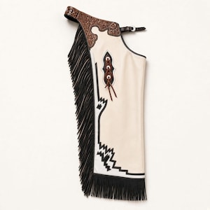 May include: A cream-coloured leather chap featuring a dark brown floral-tooled leather waistband with a buckle closure. The leg panel displays black geometric trim, a decorative lace-up detail, and long black fringe along the outer side and bottom.