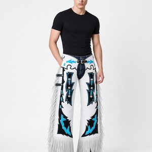 May include: White leather chaps featuring black and bright blue decorative overlays, silver-toned metal conchos, and long fringe along the outer legs. The chaps include adjustable black side straps with buckles and a front belt closure.