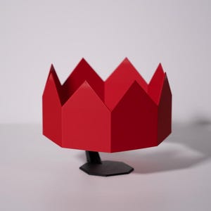 May include: A striking red, geometric decorative object, crown-shaped. It is mounted on a black, hexagonal base with a slender stem. The object is set against a plain white background. The overall design is modern and minimalist.