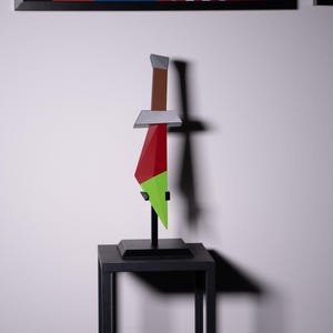 Könnte beinhalten: Eine geometrische Skulptur eines Dolches mit braunem Griff, silbernem Parierstück und roter und grüner Klinge. Der Dolch wird auf einem schwarzen Ständer vor einer weißen Wand mit abstrakter Kunst im Hintergrund ausgestellt.