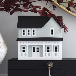 May include: A miniature house model with a light blue exterior and a black roof. The house features white trim, windows, and a porch. It sits atop a black box with a brass handle, next to a white vase and burgundy branches.