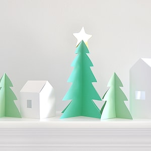 Pode incluir: Uma cena minimalista de uma vila de Natal com árvores e casas de papel. A árvore central é verde vibrante, encimada por uma estrela dourada. Duas árvores verdes menores e duas casas brancas com janelas recortadas completam a exibição festiva.
