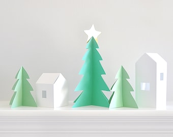 Op de afbeelding: Een minimalistische kerstdorpscène met papieren bomen en huizen. De centrale boom is een levendig groen, bekroond met een gouden ster. Twee kleinere groene bomen en twee witte huizen met uitgesneden ramen maken de feestelijke weergave compleet.