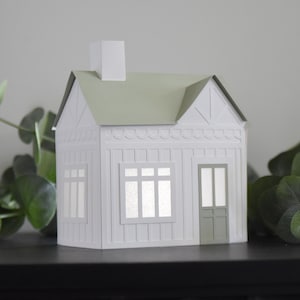 Puede incluir: Una casa de papel blanco con un techo verde y una chimenea blanca. La casa tiene dos ventanas y una puerta. La casa está sentada sobre una superficie negra.