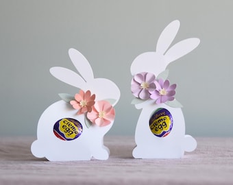 May include: Two white paper bunny shaped boxes with cutouts for candy. The bunnies are decorated with pink and purple paper flowers. The boxes contain Cadbury Creme Eggs.