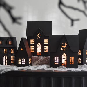 May include: A collection of five black paper houses with cut-out windows and doors, illuminated from within by a warm orange light. The structures feature intricate details including a crescent moon, stars, and flying bats, creating a glowing display.