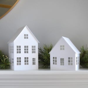 以下が含まれることがあります： 白い紙でできた家が2つ、切り抜かれた窓があり、白い暖炉の上に置かれています。両側に緑の松の枝があります。